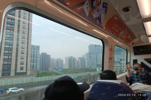 Eine Gruppe von Menschen sitzt auf einem Zugdach und schaut aus dem Fenster auf Fahrzeuge, einen Zaun, Bäume, Gebäude und den Himmel, wobei eine Person rechts ein Mobiltelefon hält.