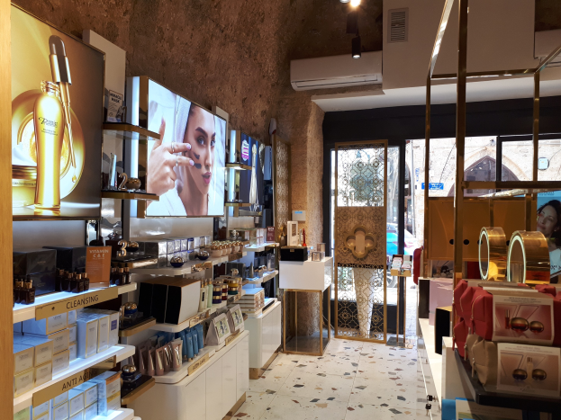 Ein hell erleuchteter Kosmetikgeschäftsinnenraum mit Regalen, die verschiedene Make-up- und Pflegemittel, Wandbildschirme, Deckenleuchten, eine Klimaanlage und Glastüren zeigen, durch die man eine Person, ein Auto und ein Gebäude sehen kann.