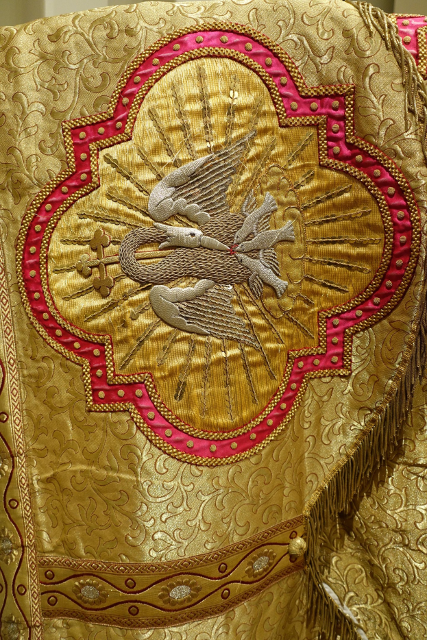 Nahaufnahme eines Priestergewandes mit aufwändiger Gold- und Rotstickerei vor einem neutralen Wandhintergrund.