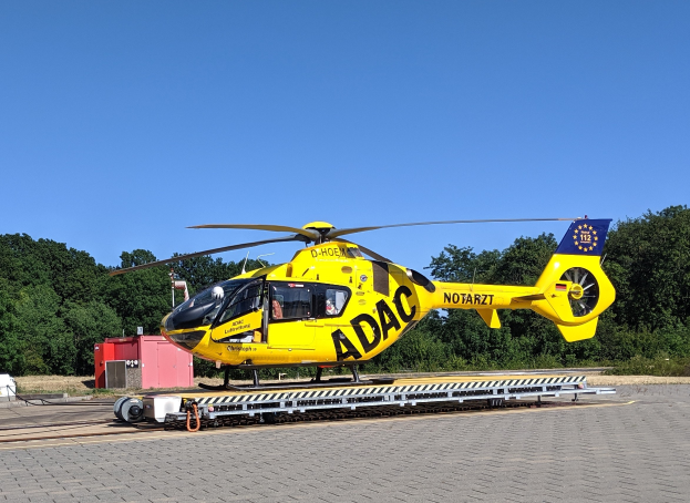 Ein gelbes Hubschrauber sitzt auf einem Flughafen-Asphalt umgeben von Bäumen und einem klaren blauen Himmel, mit verschiedenen Gegenständen in der Nähe des Hubschraubers.