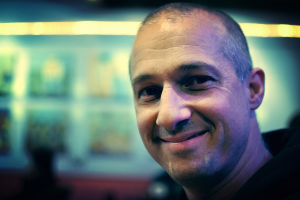 Ein Mann in einem schwarzen Kleid lacht vor einem unscharfen, hellen Hintergrund.