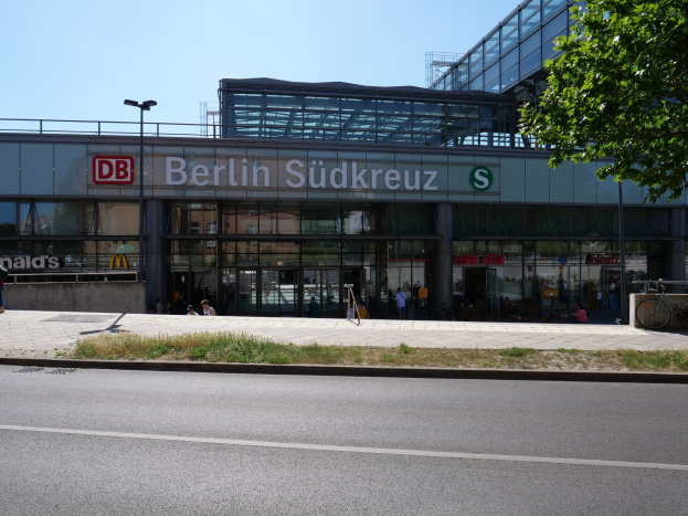 Ein großes Glaswand-Bahnhof mit der Aufschrift "Berlin Südkreuz" in Berlin, Deutschland, mit Straßenlaternen, Laternen, Fahrzeugen, Fußgängern, Fahrrädern, Bäumen und einem klaren blauen Himmel.