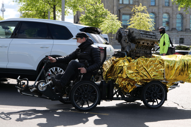 Ein Mann im Rollstuhl mit einem großen Motor am Rücken, umgeben von Fahrzeugen auf einer Straße mit Bäumen, Gebäuden und einem klaren blauen Himmel im Hintergrund, der eine schwarze Jacke und eine Kappe trägt und ein Objekt hält.