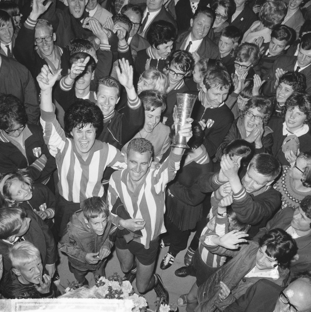 Schwarzes Bild einer Gruppe von Menschen, die mit einem Pokal feiern, Blumen unten und eine Menge im Hintergrund.