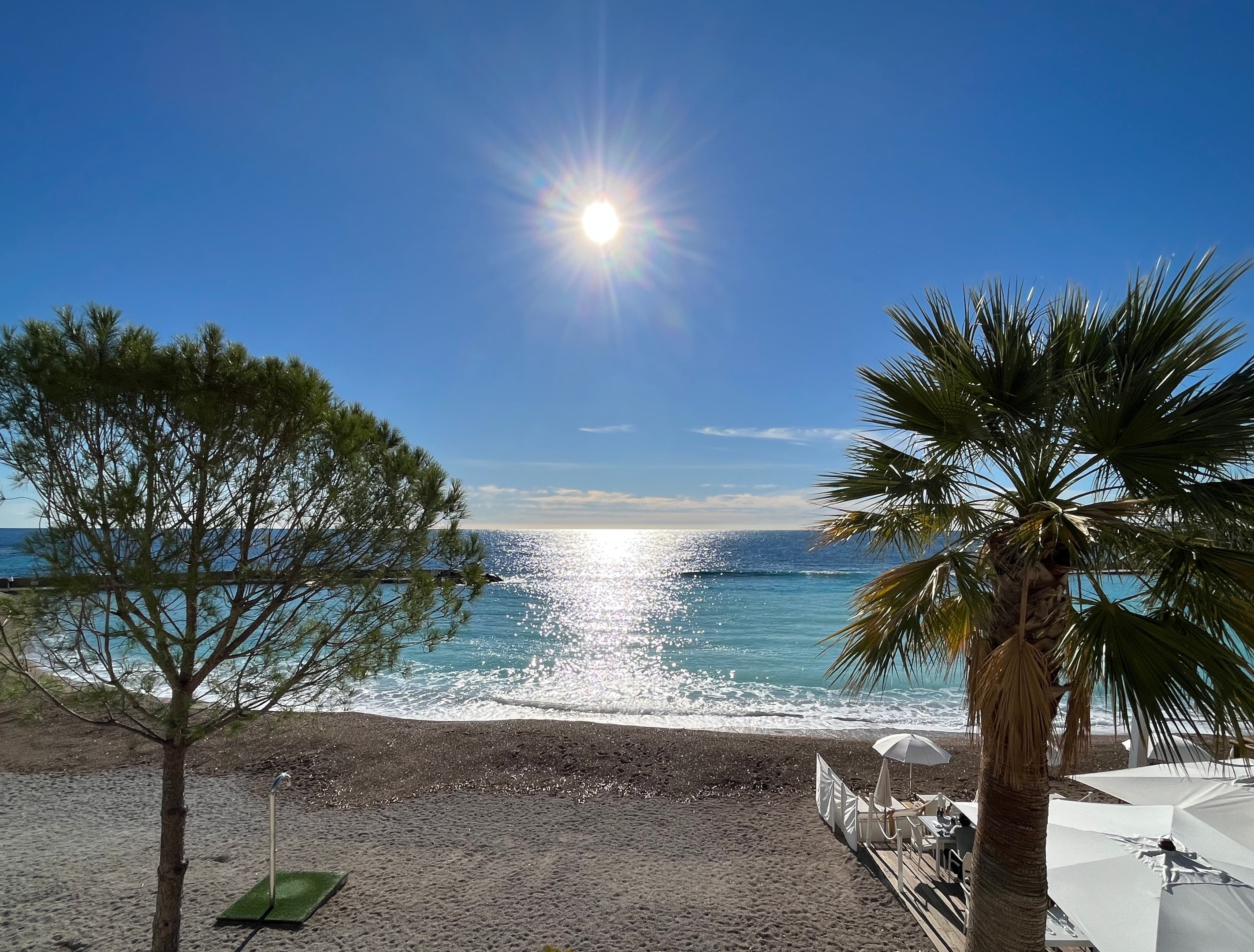 Ein Strand mit Palmen und Sonnenschirmen auf dem Sand, umgeben von saftigem Grün und Pflanzen unter einem hellblauen und weißen Himmel mit der Sonne im Hintergrund.