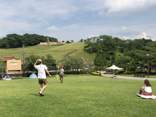 Gruppe von Menschen, die Badminton in einem Park spielt, mit einem Mann, der einen Schläger hält, anderen, die auf dem Gras sitzen, und Zelten, Polen, Gebäuden, Bäumen, Hügeln und Wolken im Hintergrund.
