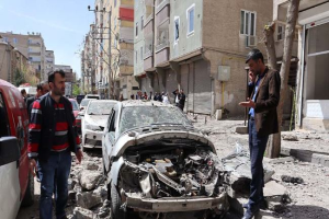 Eine Gruppe von Menschen steht um ein beschädigtes Auto auf einer Straße herum, umgeben von Gebäuden, Bäumen und Strommasten, mit einem Himmel im Hintergrund, der eine Zerstörung zeigt, die wahrscheinlich durch einen Luftangriff in Aleppo, Syrien, verursacht wurde.