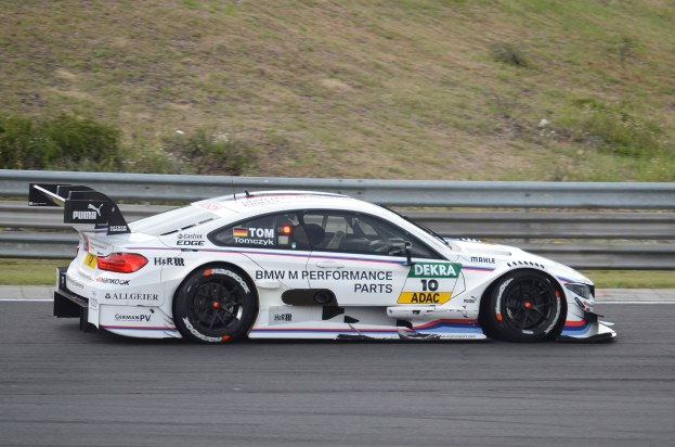 BMW M4 DTM-Rennwagen auf einer Rennstrecke mit Text auf der Karosserie, umgeben von grünem Gras und einer Begrenzung im Hintergrund.