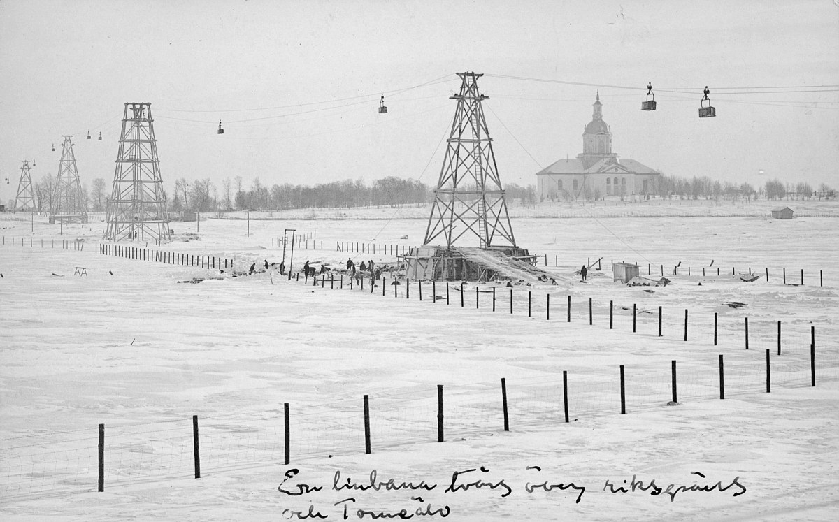 Schwarze-Weiß-Foto eines Skilifts in einer verschneiten Wiese mit Stützpfählen, Überseilbahn, Bäumen und einem Gebäude im Hintergrund, mit Text unten.