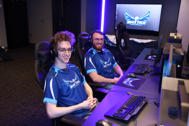 Zwei Männer mit Headsets lächeln, während sie an einem Schreibtisch mit Computerbildschirmen, Tastaturen und anderem Equipment sitzen und scheinbar ein Videospiel in einem Raum mit einem Bildschirm und Lichtern im Hintergrund spielen.