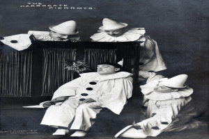 Ein Schwarz-Weiß-Foto einer Gruppe von Clowns, die auf einer Bühne sitzen, wobei eine Person ein Buch hält und Vorhänge im Hintergrund zu sehen sind.