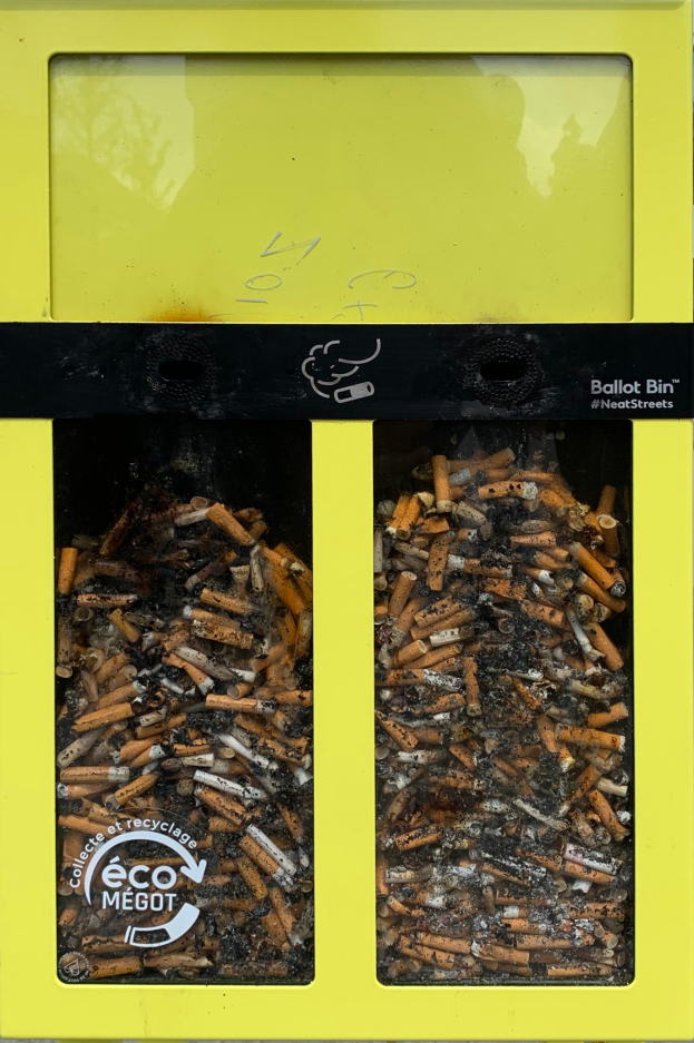 Ein leuchtend gelber Recycling-Eimer voller Zigarettenstummeln auf einem Gehweg, beschriftet für Zigarettenabfall.