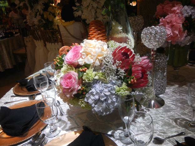 Ein formeller Esstisch mit Tellern, Besteck, Gläsern, Servietten und einem Blumenstrauß in der Mitte, umgeben von Stühlen, Tischen und sitzenden Gästen im Hintergrund.