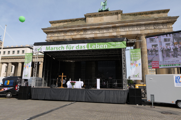 Eine Bühne vor dem Brandenburger Tor in Berlin, Deutschland, mit einem Tisch mit einer Vase, einem Kreuzsymbol und anderen Gegenständen sowie Bannern, Lautsprechern und Fahrzeugen auf der Straße.