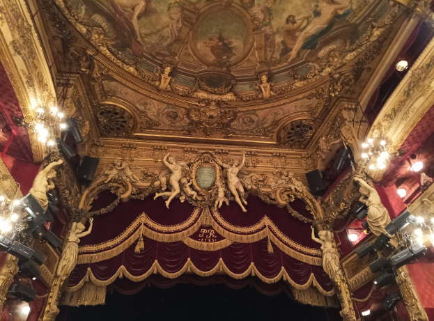 Theaterinnenraum mit roter Gardine, Kronleuchtern, Skulpturen, Lichtern und einer bemalten Decke, mit einer Bühne und einer kleinen Gruppe von Menschen unten.