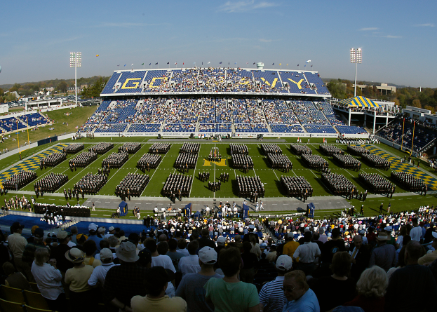 Eine große Menschenmenge in einem Stadion bei einem Fußballspiel, wobei einige stehen und andere sitzen, auf einem grünen Feld unter einem klaren blauen Himmel, mit Laternenmasten, Bäumen und Gebäuden im Hintergrund.
