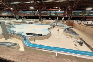 Ein großes Indoor-Wasserpark mit zahlreichen Wasserrutschen, Stangen, Texttafeln, Deckenleuchten und Wandgemälde, das wie ein Minigolfplatz gestaltet ist.