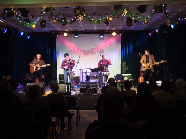 Eine Gruppe von Menschen, die auf der Bühne mit Musikinstrumenten, Mikrofonen und Ständern spielen, während das Publikum in Stühlen im Vordergrund unter festlicher Beleuchtung und Dekoration sitzt.