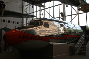 Ein Flugzeug von American Airlines wird in einem Museum ausgestellt, mit Menschen auf dem Boden und Treppen, die auf der rechten Seite zum Flugzeug führen, sowie einem Gebäude, Bäumen und einem klaren blauen Himmel im Hintergrund.