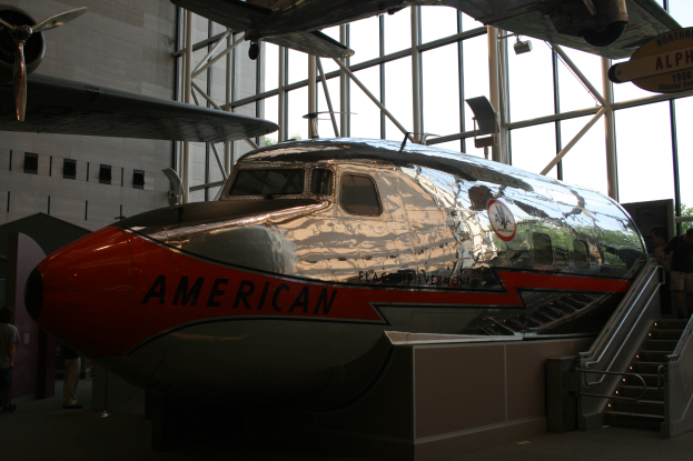 Ein Flugzeug von American Airlines wird in einem Museum ausgestellt, mit Menschen auf dem Boden und Treppen, die auf der rechten Seite zum Flugzeug führen, sowie einem Gebäude, Bäumen und einem klaren blauen Himmel im Hintergrund.