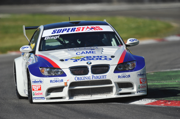 Ein BMW M3 GT3 Rennauto fährt auf einer unscharfen Rennstrecke mit Gras im Hintergrund.