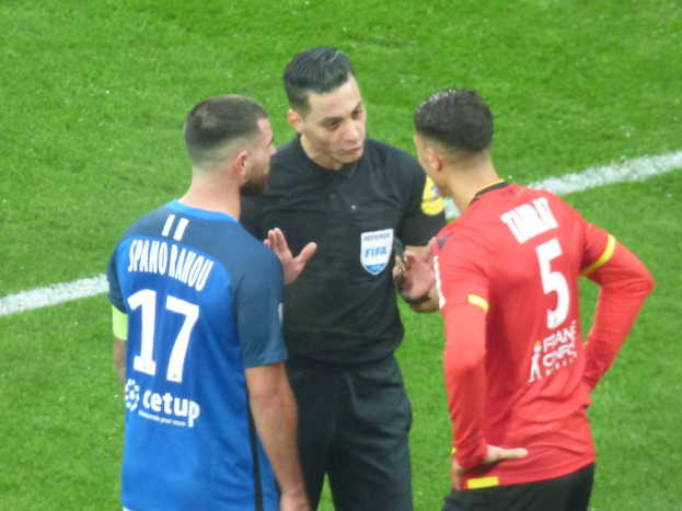 Schiedsrichter spricht mit zwei Männern auf einem Fußballfeld; einer trägt ein schwarzes Hemd und Hosen, der andere ein blaues und rotes Hemd mit Text und Brille.