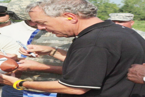 Ein Mann in einem schwarzen Hemd unterschreibt einen Fussball, während er von einer Gruppe von Menschen umgeben ist, die Kappen und Schutzbrillen tragen. Im Hintergrund sind Bäume und ein klarer blauer Himmel zu sehen.