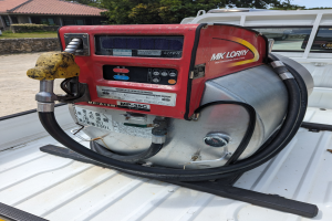 Ein Lastwagen mit einer Tankröhre auf der Ladefläche, umgeben von Bäumen, einem Haus und einem klaren blauen Himmel, ausgestattet mit einem Motor und einer Zapfsäule zum Betanken von Fahrzeugen.