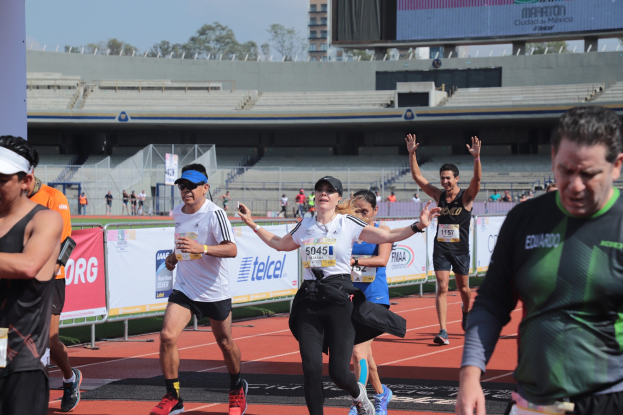 Gruppe von Menschen, die auf einer Outdoor-Sportanlage laufen, mit Bannern, einer Anzeigetafel, einem Gebäude, Bäumen und einer bewölkten Himmel.