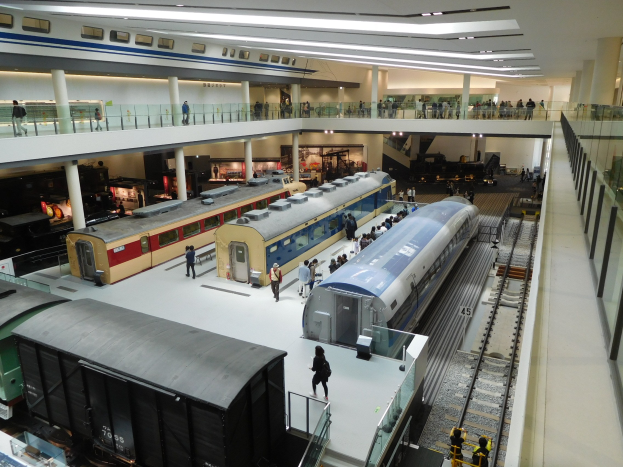 Eine geräumige Museumsausstellung, die verschiedene Züge in unterschiedlichen Größen und Farben zeigt, mit Menschen, die vom Boden, Geländern und Säulen aus beobachten.