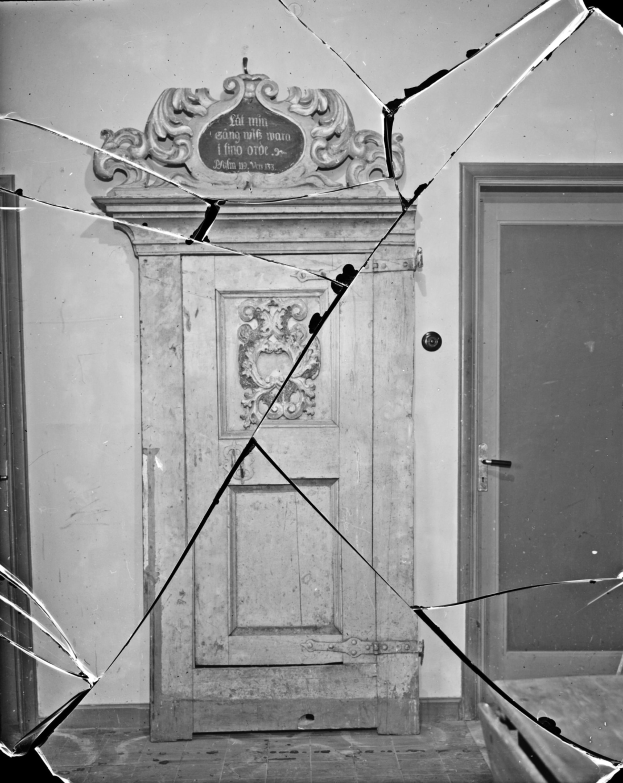 Schwarzes und weißes Foto einer kaputten Tür in einem Raum mit einer Wand im Hintergrund, die Tür in einem desolaten Zustand mit verstreuten Glasscherben.