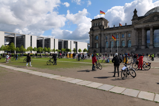 Menschen auf Fahrrädern vor dem Reichstaggebäude in Berlin, Deutschland, mit Flaggen, Säulen, bewölktem Himmel, grasigem Boden und Bäumen im Hintergrund.