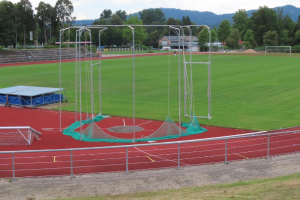 Ein Fußballfeld mit einem Netz, umgeben von einem Metallzaun, Gras, Bäumen, Häusern, Pfosten und einer Treppe, unter einem bewölkten Himmel, mit einem Trampolin in der Mitte.