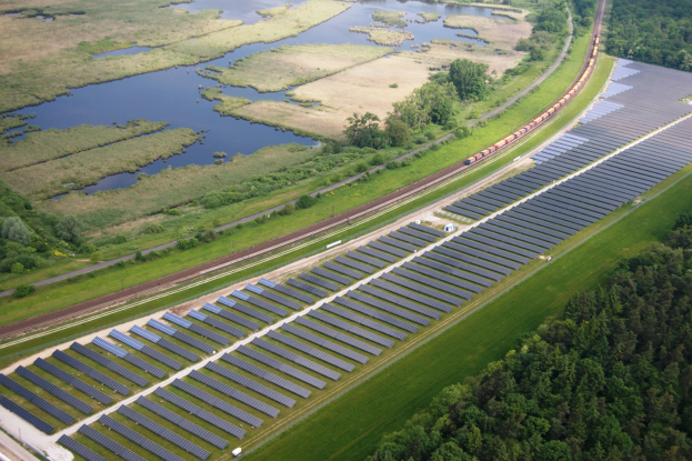 Luftaufnahme einer Solar-Farm mit Panelen auf einem grasbewachsenen Feld, umgeben von Bäumen und Wasser, mit einem Zug auf einer nahen Schiene.
