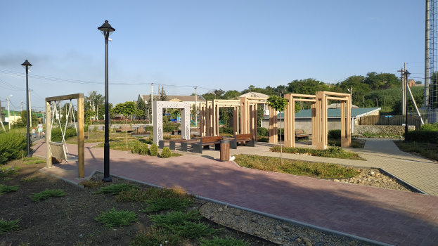 Ein Parkweg mit Holzpfählen, Bänken, Pflanzen, Gras, Steinen, Straßenlaternen, Strommasten mit Kabeln, Bäumen, Gebäuden und einem Turm unter einem klaren blauen Himmel.