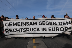 Eine Gruppe von Menschen auf der Straße mit einem Banner, auf dem 'Gemeinsam gegen den Rechtsruck in Europa' steht, vor Straßenschildern, Laternen, Strommasten, Kabeln, Gebäuden und einem bewölkten Himmel.