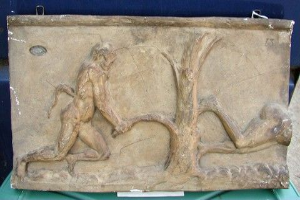 Eine Steintafel mit einer Reliefskulptur eines Mannes und einer Frau, der Mann mit ausgestrecktem Arm und die Frau mit gebeugtem Arm, vor einem blauen Hintergrund mit einer grünen Oberfläche.