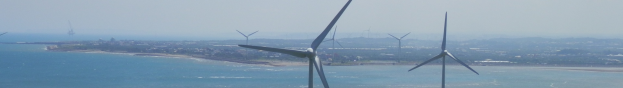Gruppe von Windkraftanlagen im Meer in der Nähe einer Stadt, mit Wasser, das gegen die Küste schwappt, Bäumen und Gebäuden im Hintergrund und einem klaren blauen Himmel darüber.