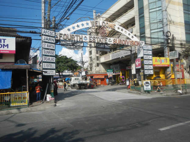 Eine belebte Stadtstraße mit Fahrzeugen, Fußgängern, Gebäuden und einem Schild mit der Aufschrift "Vierter Stand" in der Mitte, unter einem bewölkten Himmel.