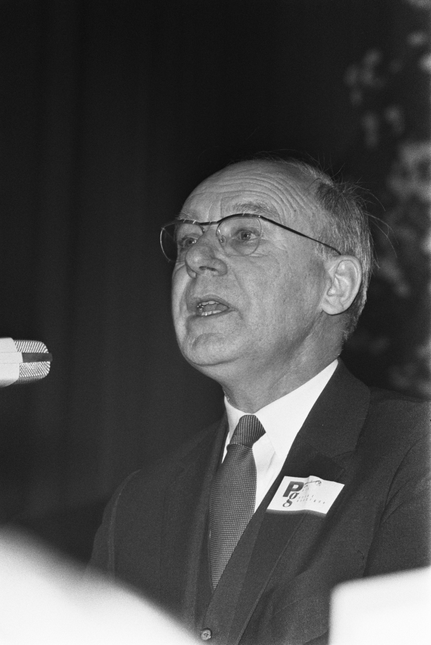 Ein Mann im Anzug und mit Krawatte spricht in ein Mikrofon, trägt eine Brille und hat einen ernsten Gesichtsausdruck, auf einem Schwarz-Weiß-Bild.