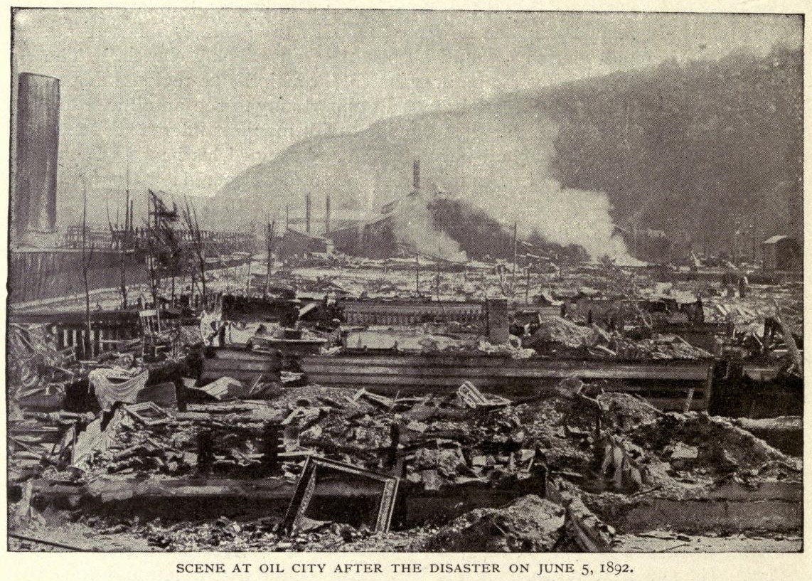 Schwarzes und weißes Foto einer Stadt nach einer Katastrophe am 5. Juni 1892, das Gebäude, Pfähle, Rauch, einen Hügel im Hintergrund und Text am unteren Rand zeigt.