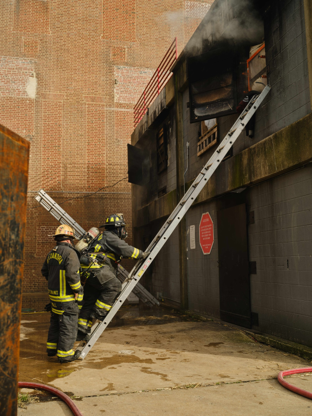 Feuerwehrleute mit Helmen klettern eine Leiter zu einem rauchgefüllten Gebäude hinauf, mit einem Metallobjekt links und Rohren am Fuß.