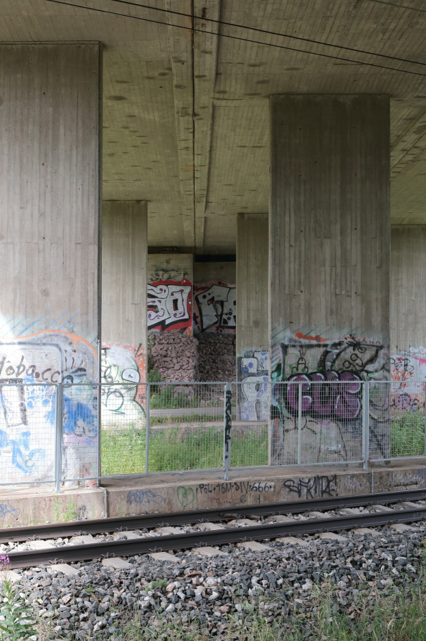 Eisenbahnschiene mit Graffiti an der Seite, umgeben von Steinen, Gras, einem Zaun, Pfeilern, Drähten und einem Dach.