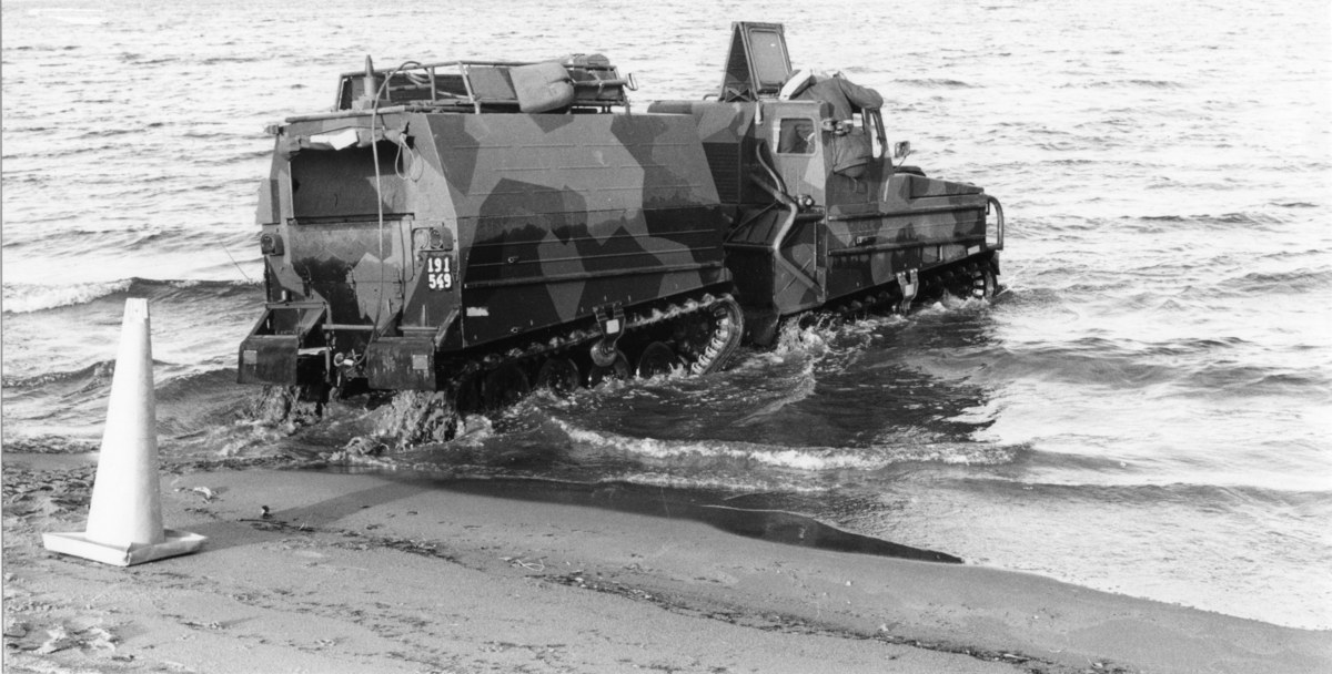 Schwarzes und weißes Foto eines Amphibienfahrzeugs, das durch Wasser fährt, mit einem Verkehrskegel links und Sand unten.