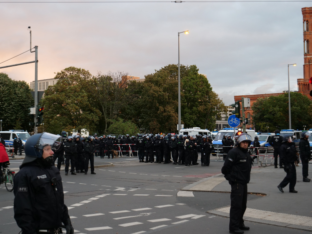 Eine Gruppe von Polizisten in Helmen, die auf einer Straße stehen, umgeben von Fahrzeugen, Verkehrsampeln, Laternenmasten, Bäumen, Gebäuden und einem bewölkten Himmel.