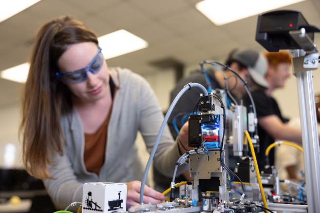 Eine Frau mit Schutzbrille, die an einem Roboter in einem Labor arbeitet, umgeben von Menschen und verschiedenen Gegenständen auf dem Tisch.