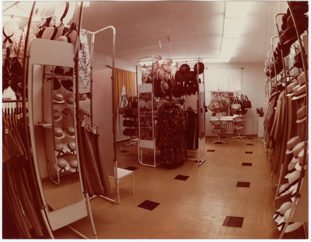 Altes Foto eines Kleiderladen-Interieurs mit Kleiderständern, Hutständern, einem Spiegel, einem Stuhl, einem Tisch mit Gegenständen, Vorhängen und Deckenleuchten.