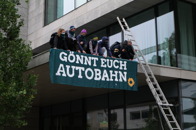 Eine Gruppe von Menschen auf einem Gebäudedach hält eine Fahne, mit einem Baum links und Glasfenstern im Hintergrund, die scheinbar protestieren, um die deutsche Regierung zu unterstützen.