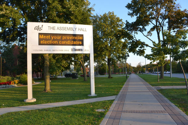 Eine Kreidetafel auf Gras neben einem Weg mit der Aufschrift "Die Versammlungshalle - Treffen Sie Ihre Landtagswahlkandidaten" mit Bäumen, Blumen, Straßenlaternen, Fahrzeugen, einem Gebäude und einem bewölkten Himmel im Hintergrund.