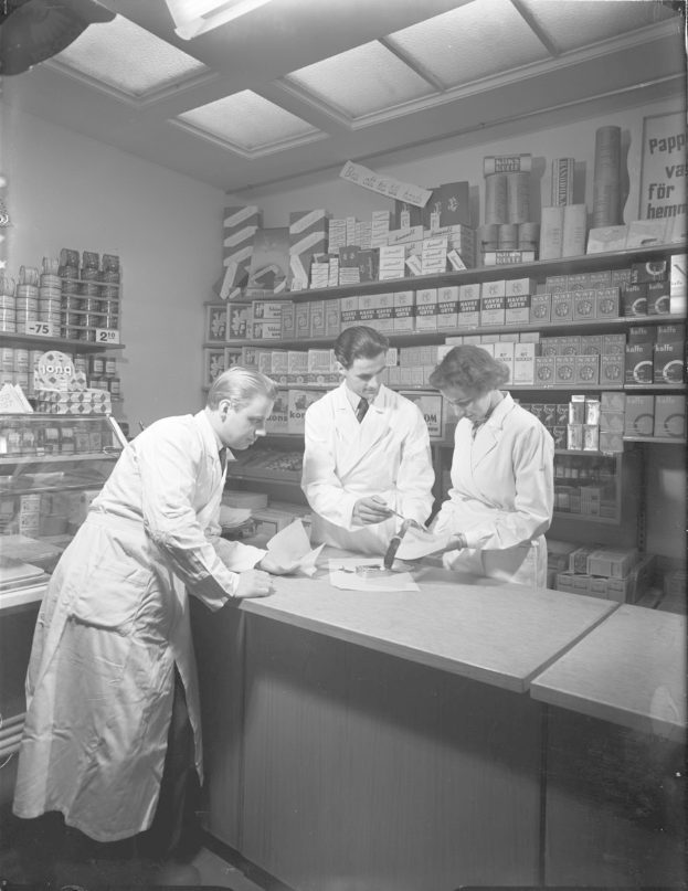 Drei Männer in weißen Kitteln an einer Apotheken-Theke mit Papieren und Gegenständen, auf einem Schwarz-Weiß-Bild mit Regalen voller Artikel im Hintergrund.